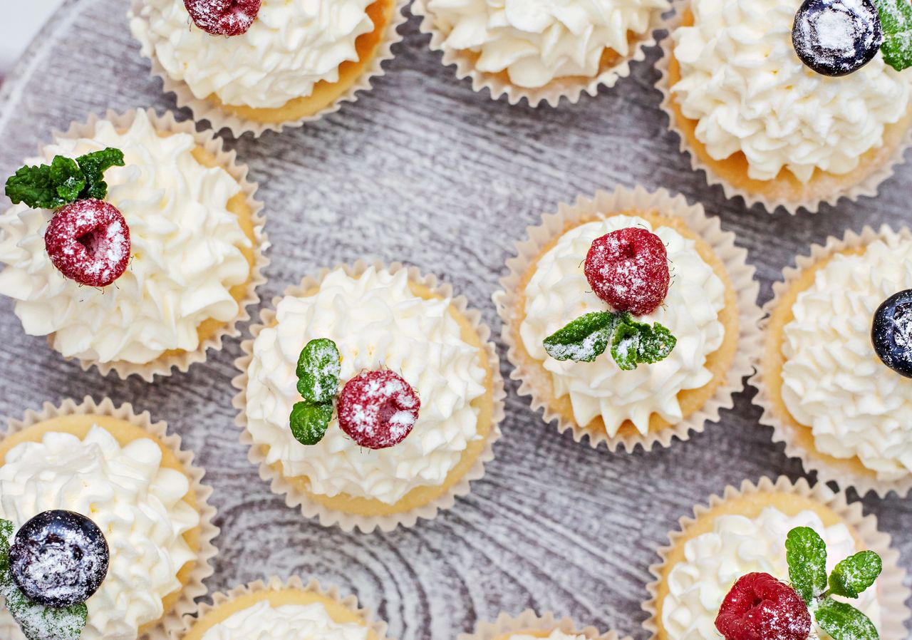 hero-img-02 A vibrant overhead shot of cupcakes topped with whipped cream, raspberries, blueberries, and mint leaves.