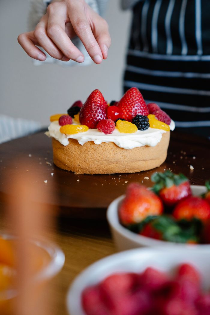 gallery-3 Close-up of a fresh fruit-topped cake being finished with a hand adding berries.